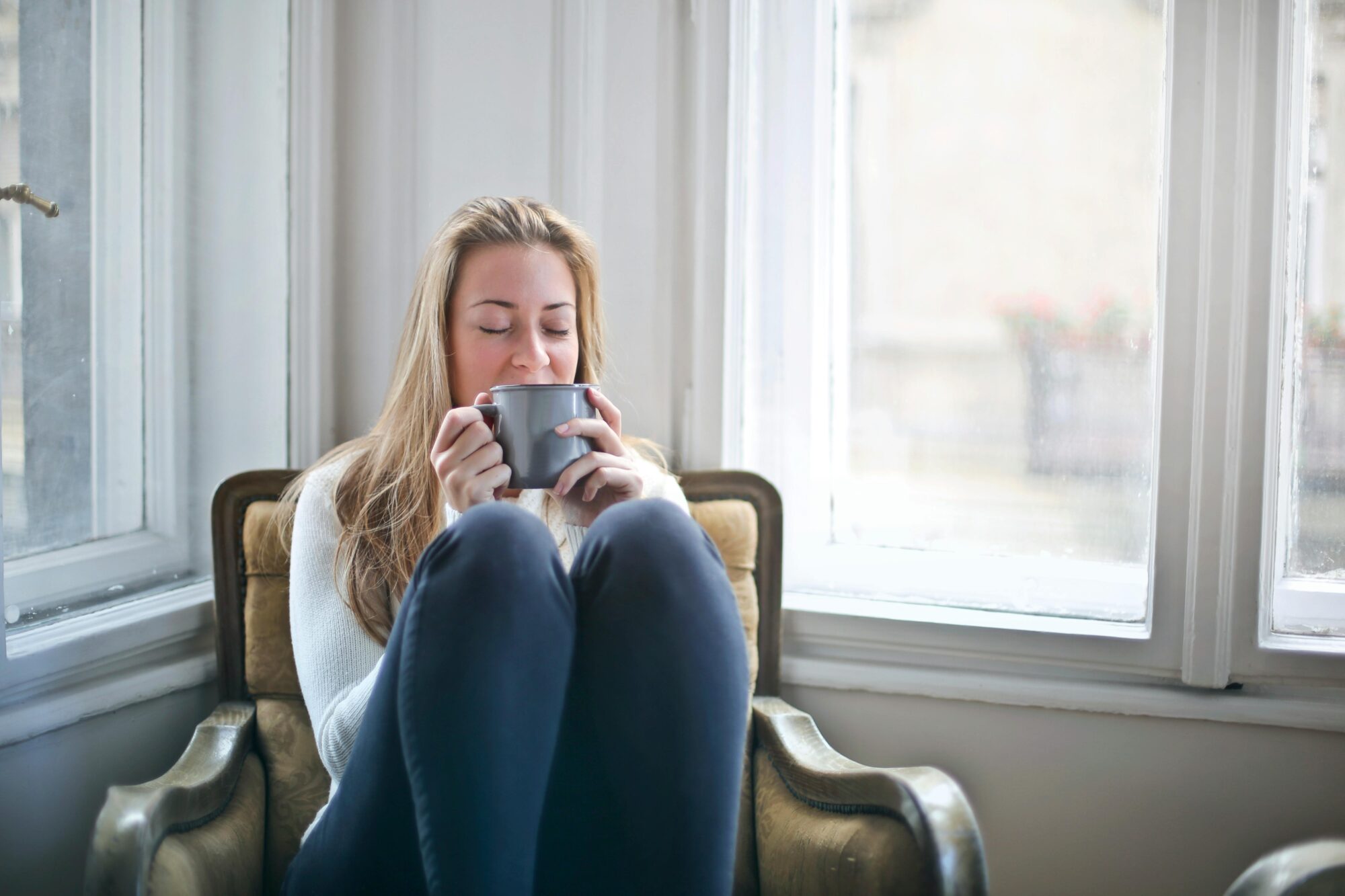 Junge Frau sitzt gem&uuml;tlich in einem Sessel am Fenster und h&auml;lt eine warme Tasse in den H&auml;nden &ndash; ein typischer Hygge-Moment voller Ruhe und Wohlbefinden.