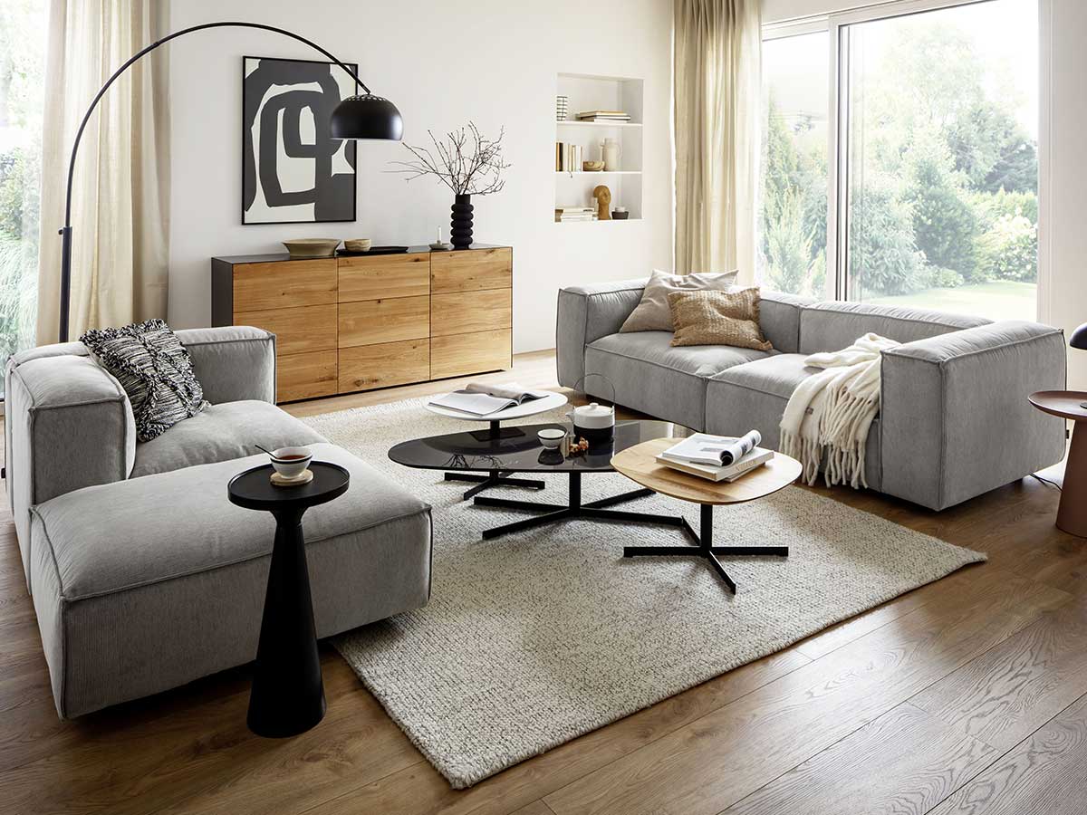 Wohnzimmer mit hellgrauen Polstermöbeln, naturfarbenem Sideboard und schwarzen Dekoartikeln
