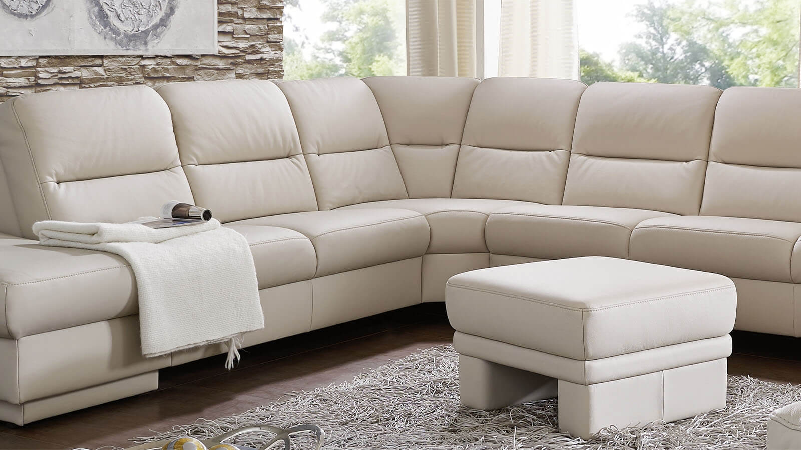 Große beige Wohnlandschaft mit großzügiger Sitzfläche, gepolsterten Rückenlehnen und passendem Hocker in einem hellen Wohnzimmer