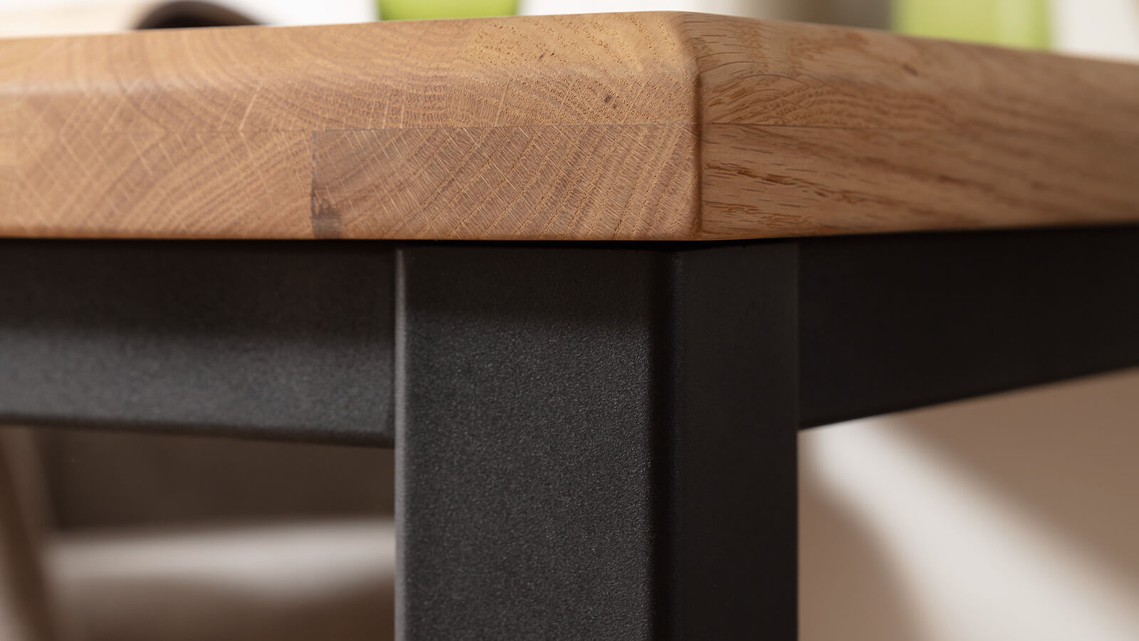Nahaufnahme der abgeschrägten Kante einer geölten Eichen-Tischplatte mit ausgeprägter Holzmaserung auf einem eckigen Gestell aus mattschwarzem Stahl im modernen Industrial-Stil