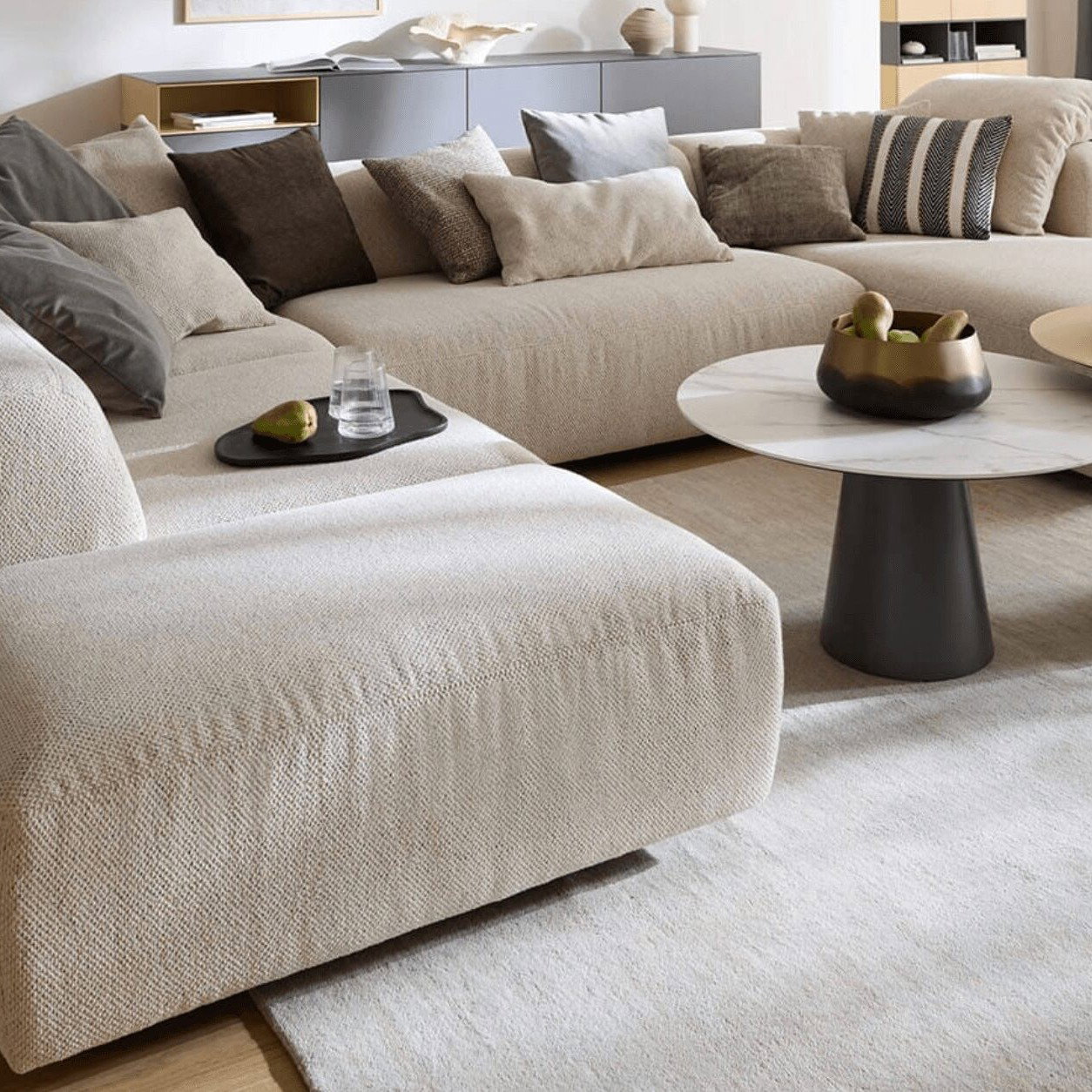 Helle Wohnlandschaft mit vielen Kissen und modernen Couchtischen in einem stilvollen Wohnzimmer.Ein großzügiges Sofa gehört zu den wichtigsten erste Wohnung Möbeln. Es bietet ausreichend Platz für den Alltag und wird schnell zum Lieblingsplatz in den eigenen vier Wänden.