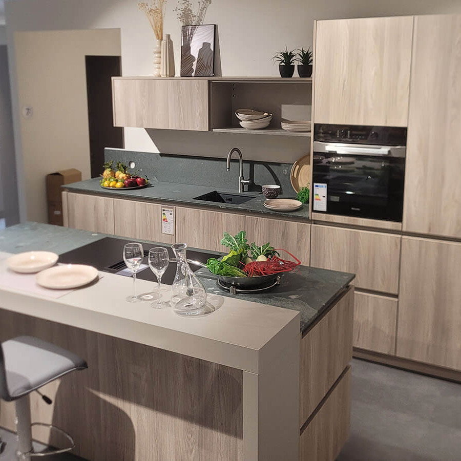 Helle Ausstellungsküche in Esche Nordic mit dunkler Arbeitsplatte, Kücheninsel, Sitzbereich und Elektrogeräten von Miele.