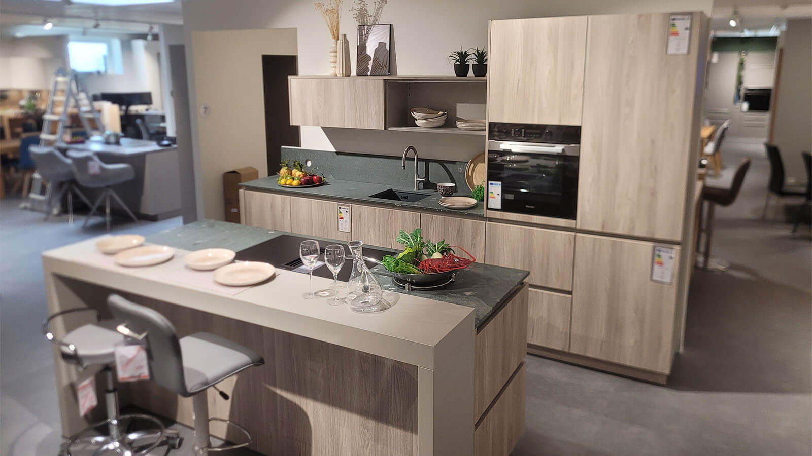Helle Ausstellungsküche in Esche Nordic mit dunkler Arbeitsplatte, Kücheninsel, Sitzbereich und Elektrogeräten von Miele.