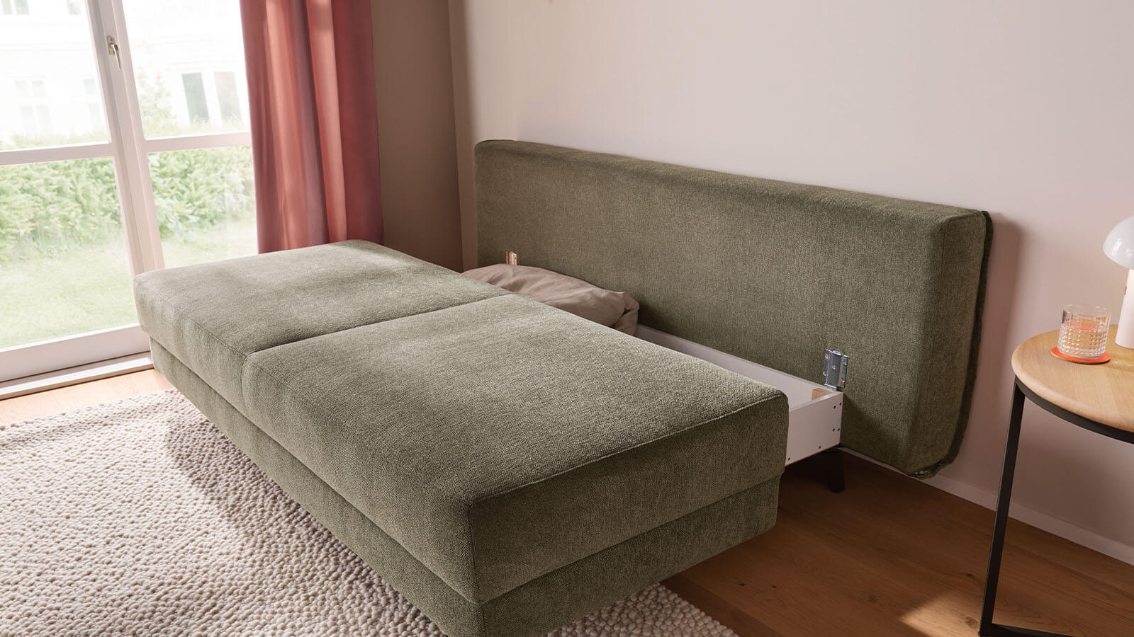 Großzügiges grünes Schlafsofa mit weichem Stoffbezug, lässiger Polsterung und gemütlichen Armlehnenkissen. Die schwarzen Metallfüße setzen einen modernen Akzent, während der integrierte Bettkasten praktischen Stauraum bietet.