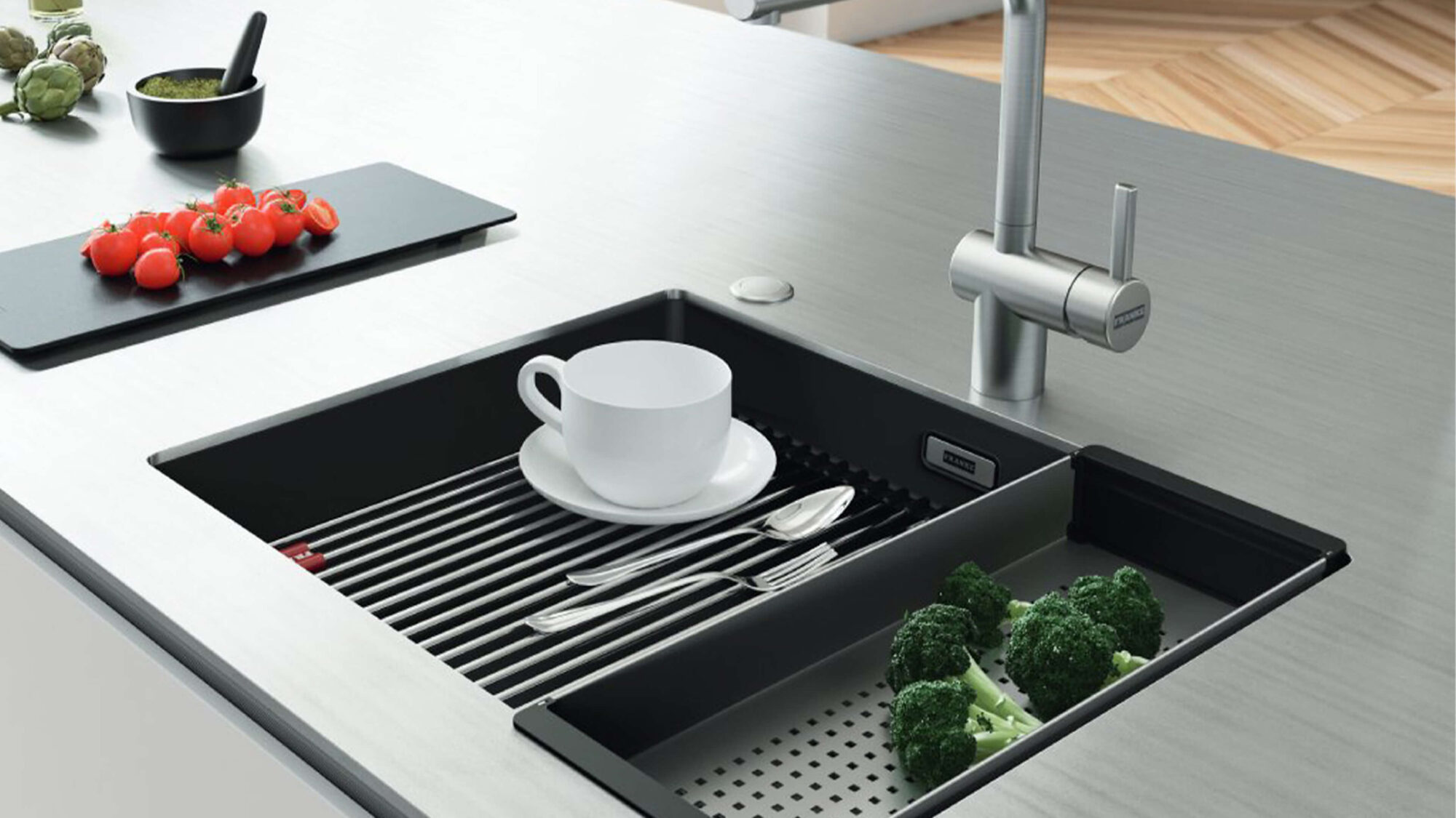 Moderne K&uuml;che mit einer gro&szlig;fl&auml;chigen hellen Edelstahl-Arbeitsplatte. In die Arbeitsfl&auml;che ist eine schwarze Sp&uuml;le mit integriertem Abtropfbereich eingesetzt. Auf der Sp&uuml;le stehen eine wei&szlig;e Tasse und Besteck, daneben liegt frisches Gem&uuml;se auf einem schwarzen Schneidebrett. Im Hintergrund ist eine Edelstahl-Armatur vor einer K&uuml;chenzeile mit Holzoberfl&auml;chen zu sehen.