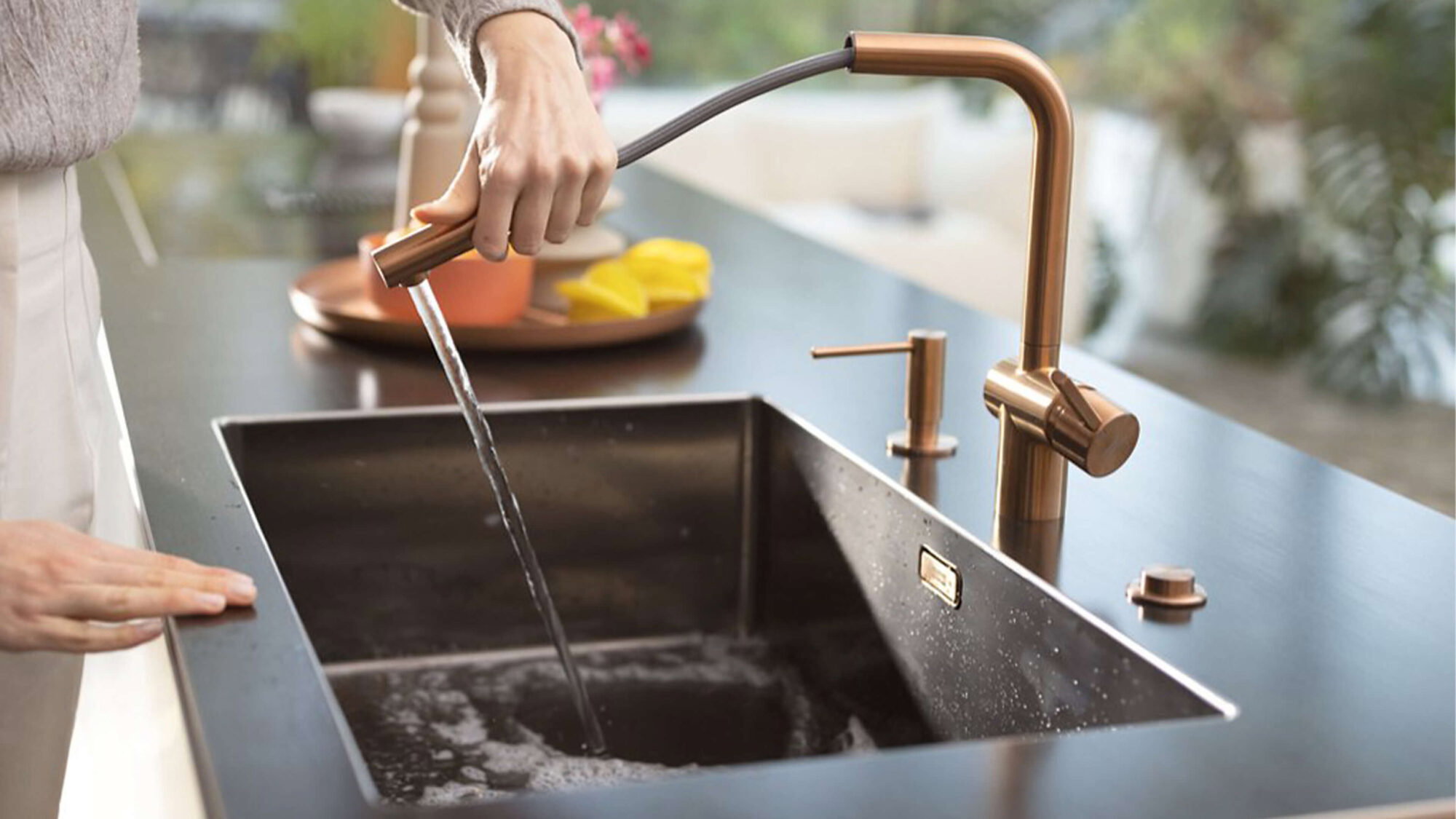 Detailansicht einer dunklen K&uuml;chenarbeitsplatte mit gro&szlig;er schwarzer Sp&uuml;le und einer gebogenen Armatur in Kupferoptik. Wasser l&auml;uft aus der Armatur in das Sp&uuml;lbecken. Eine Person bedient die Armatur mit der Hand. Im Hintergrund sind K&uuml;chenutensilien und frisches Obst in warmem Tageslicht zu sehen.